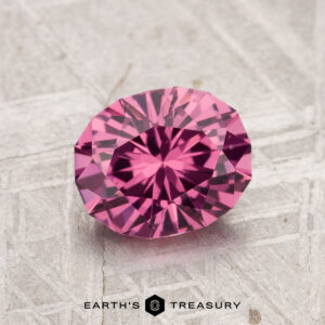 A round, faceted 1.52-carat Tanga Garnet with a pink hue is displayed on a textured, light-colored surface, with the text "EARTH'S TREASURY" below.