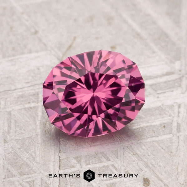 1.52-Carat Tanga Garnet A round, faceted 1.52-carat Tanga Garnet with a pink hue is displayed on a textured, light-colored surface, with the text "EARTH'S TREASURY" below.