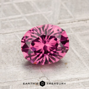 A 1.51-carat Tanga Garnet, round and faceted in pink, rests on a gray textured surface with the Earth’s Treasury logo beneath it.