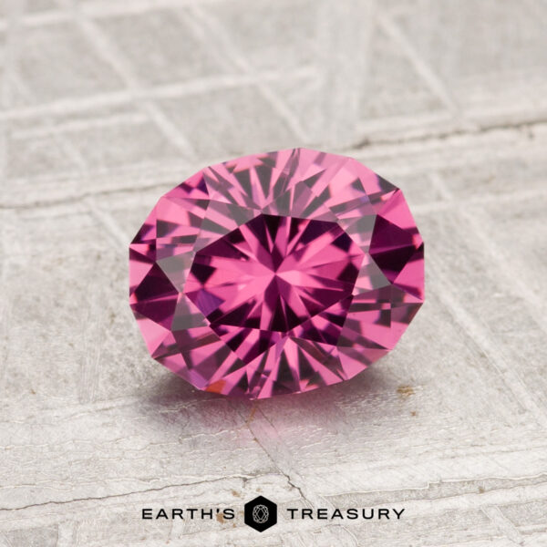 1.51-Carat Tanga Garnet A 1.51-carat Tanga Garnet, round and faceted in pink, rests on a gray textured surface with the Earth’s Treasury logo beneath it.