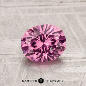 A 1.33-carat Tanga Garnet, round brilliant-cut and pink, is shown on a textured light gray surface with the Earth’s Treasury logo and text beneath it.