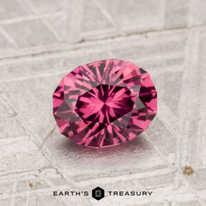 The 1.19-Carat Tanga Garnet, a faceted oval-cut pink gemstone, is displayed on a textured light gray surface with the Earth's Treasury logo at the bottom.