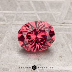 A 1.19-carat Tanga Garnet, an oval faceted pink gemstone, is showcased on a textured surface with the words "EARTH'S TREASURY" at the bottom.