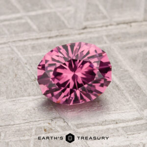 A 1.19-carat faceted oval Tanga Garnet rests on a textured metallic surface, with "EARTH'S TREASURY" displayed at the bottom of the image.