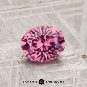 A 1.12-carat oval Tanga Garnet is displayed on a textured light gray surface, with "EARTH'S TREASURY" at the bottom.