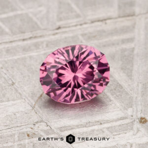 A 0.88-carat Tanga Garnet, faceted in an oval cut and displaying a pink hue, rests on a textured metallic surface with "Earth's Treasury" at the bottom.