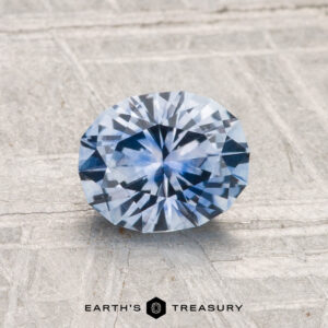 A 1.27-carat Montana Sapphire (Heated) with an oval cut is displayed on a textured light gray surface, with "EARTH’S TREASURY" written at the bottom.