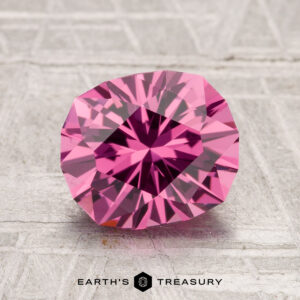 A 2.41-carat Tanga Garnet gemstone with a pink hue is displayed on a textured gray surface, featuring the text "EARTH'S TREASURY" at the bottom.