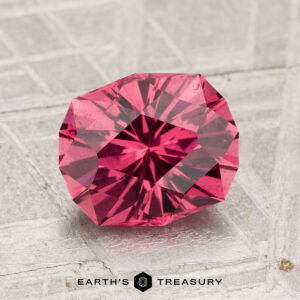 The 2.38-Carat Tanga Garnet, a faceted deep pink gemstone, is showcased on a textured light gray surface with “EARTH’S TREASURY” written below.