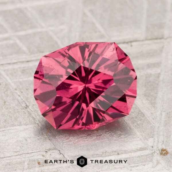 2.38-Carat Tanga Garnet The 2.38-Carat Tanga Garnet, a faceted deep pink gemstone, is showcased on a textured light gray surface with “EARTH’S TREASURY” written below.