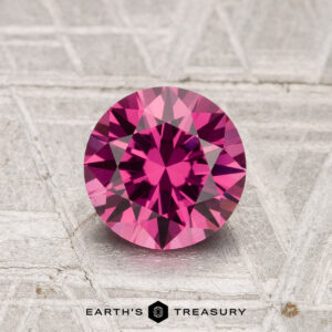 A 1.34-carat Tanga Garnet, cut in a faceted round shape, is displayed on a textured gray surface with the "Earth's Treasury" logo at the bottom.