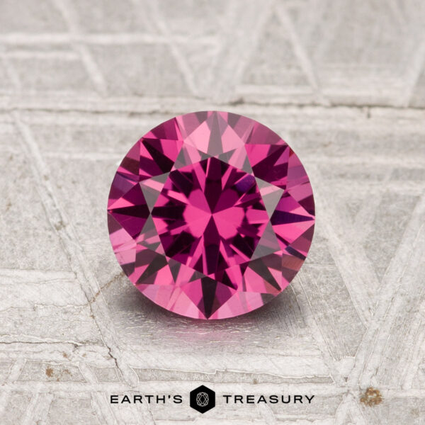 1.34-Carat Tanga Garnet A 1.34-carat Tanga Garnet, cut in a faceted round shape, is displayed on a textured gray surface with the "Earth's Treasury" logo at the bottom.