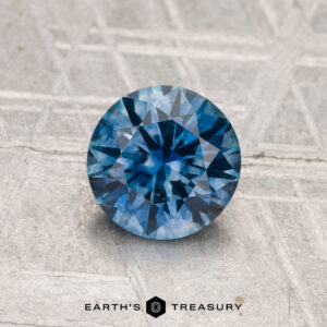 A 1.25-carat heated Montana sapphire, round brilliant-cut, is displayed on a textured gray surface with "EARTH'S TREASURY" at the bottom.