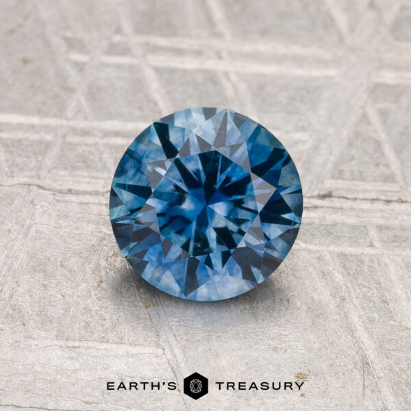1.25-Carat Montana Sapphire (Heated) A 1.25-carat heated Montana sapphire, round brilliant-cut, is displayed on a textured gray surface with "EARTH'S TREASURY" at the bottom.