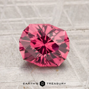 A 1.82-carat Tanga Garnet, round and faceted with a vivid pink hue, rests on a textured silvery surface, accompanied by the "Earth's Treasury" text below.