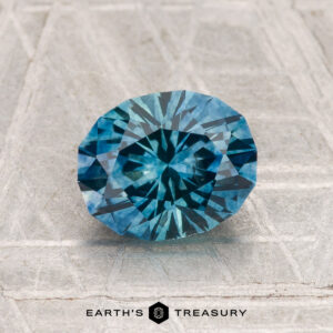 A 1.81-carat heated Montana sapphire, round brilliant-cut and blue, is shown on a textured gray surface with "EARTH'S TREASURY" displayed at the bottom.