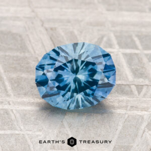 A 1.62-carat faceted blue Montana Sapphire (Heated) is displayed on a textured grey surface with the "Earth's Treasury" logo at the bottom.