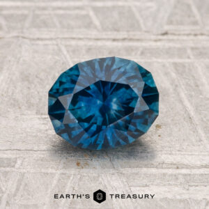 A 1.55-carat heated Montana sapphire, faceted oval and blue, is displayed on a textured light gray surface with "EARTH'S TREASURY" printed below.