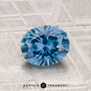 A round, faceted blue 1.52-carat Montana sapphire (heated) rests on a textured surface with "Earth’s Treasury" displayed at the bottom of the image.