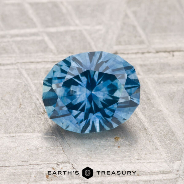 1.52-Carat Montana Sapphire (Heated) A round, faceted blue 1.52-carat Montana sapphire (heated) rests on a textured surface with "Earth’s Treasury" displayed at the bottom of the image.