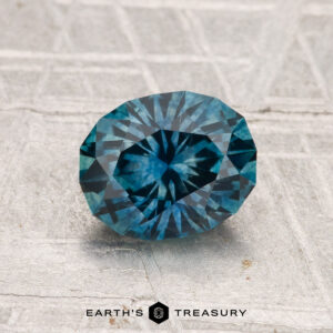 A 1.49-carat heated Montana sapphire, oval-shaped and faceted, sits on a textured gray surface with "EARTH'S TREASURY" displayed below.