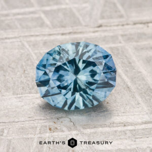 A round, faceted 1.36-carat Montana Sapphire (heated) with a blue hue is displayed on a textured, light background featuring "Earth's Treasury" at the bottom.