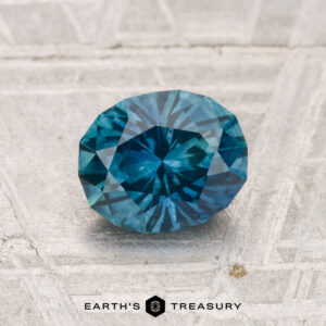 A 1.28-carat heated Montana sapphire, cut in a faceted oval shape, is displayed on a light, textured surface with "EARTH'S TREASURY" at the bottom.