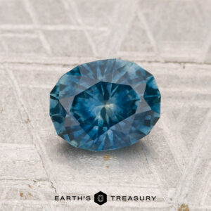 A 1.27-carat heated Montana sapphire, oval-cut and blue, is showcased on a light gray textured surface with the Earth’s Treasury logo at the bottom.