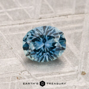 A 1.25-carat heated Montana sapphire with intricate facets rests on a textured metallic surface; the "EARTH'S TREASURY" logo is visible at the bottom.