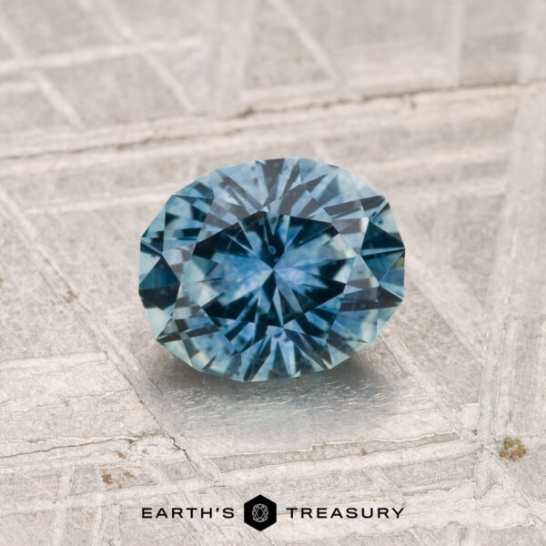1.25-Carat Montana Sapphire (Heated) A 1.25-carat heated Montana sapphire with intricate facets rests on a textured metallic surface; the "EARTH'S TREASURY" logo is visible at the bottom.