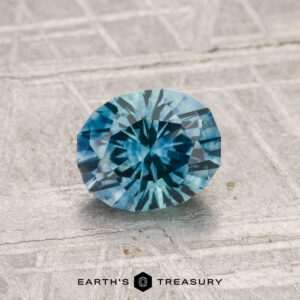 A 1.22-carat heated Montana sapphire with a blue-green hue is displayed on a textured light gray surface, featuring the text "EARTH'S TREASURY" at the bottom.