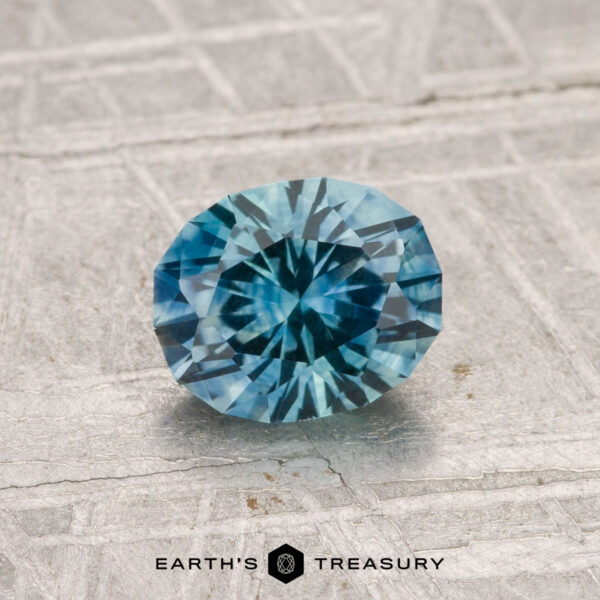 1.22-Carat Montana Sapphire (Heated) A 1.22-carat heated Montana sapphire with a blue-green hue is displayed on a textured light gray surface, featuring the text "EARTH'S TREASURY" at the bottom.