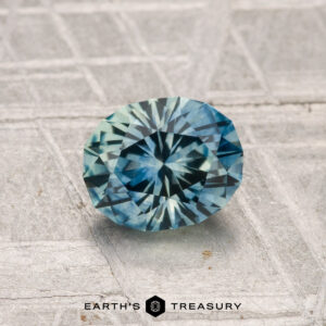 A 1.20-carat heated Montana sapphire, cut in a faceted oval and displaying blue-green hues, rests on a textured gray surface with "EARTH'S TREASURY" shown at the bottom.