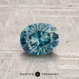 A 1.19-carat heated Montana sapphire in blue-green hues is displayed on a textured grey surface with the Earth’s Treasury logo at the bottom.