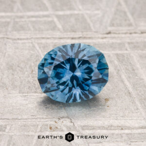 A faceted oval 1.18-carat heated Montana sapphire rests on a textured light gray surface, with the text "EARTH'S TREASURY" at the bottom.
