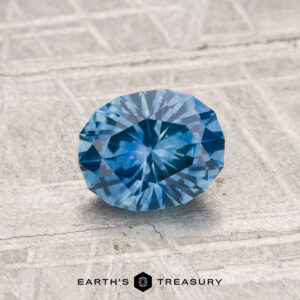 A 1.14-carat heated Montana sapphire with an oval cut displayed on a textured, light surface featuring the Earth's Treasury logo at the bottom.