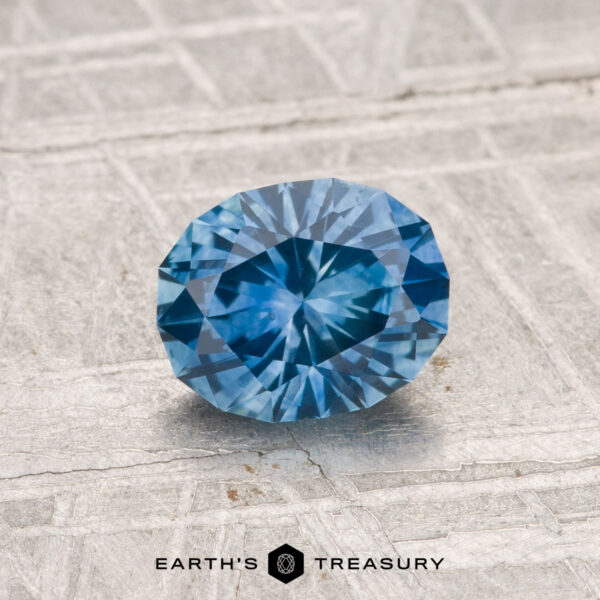1.14-Carat Montana Sapphire (Heated) A 1.14-carat heated Montana sapphire with an oval cut displayed on a textured, light surface featuring the Earth's Treasury logo at the bottom.
