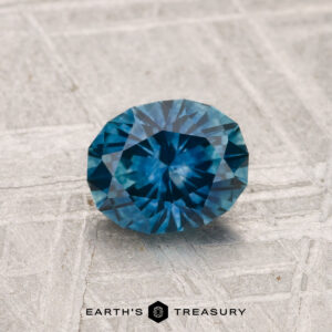 A round, faceted 1.08-carat heated Montana sapphire with a blue hue rests on a textured, light background, with "Earth's Treasury" written below.