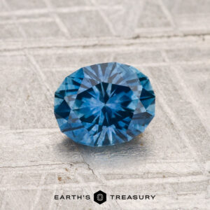 A round, faceted 1.05-carat heated Montana sapphire with a blue hue is displayed on a textured, light-colored surface featuring the text "EARTH'S TREASURY" at the bottom.