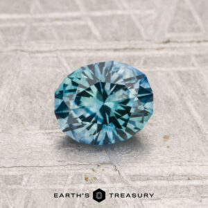 A 1.04-carat heated Montana Sapphire, featuring a faceted oval blue-green gem, is shown on a textured light gray surface with "Earth's Treasury" at the bottom.