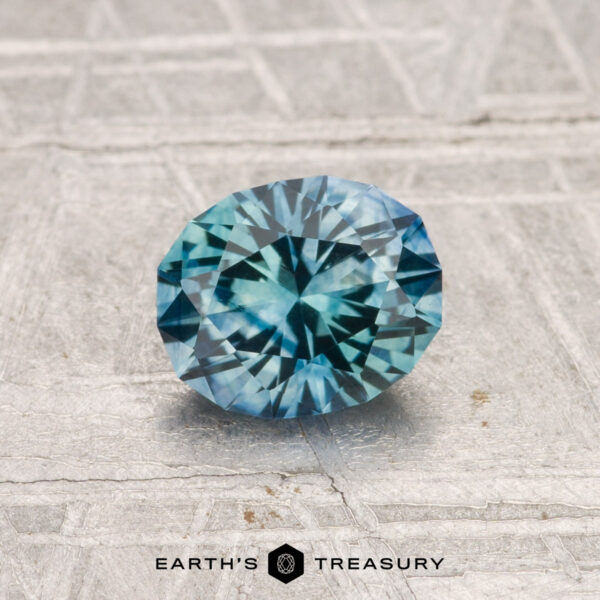 1.04-Carat Montana Sapphire (Heated) A 1.04-carat heated Montana Sapphire, featuring a faceted oval blue-green gem, is shown on a textured light gray surface with "Earth's Treasury" at the bottom.