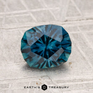 A 1.66-carat heated Montana sapphire, deep blue and faceted, is displayed on a textured light gray surface with "EARTH'S TREASURY" at the bottom.