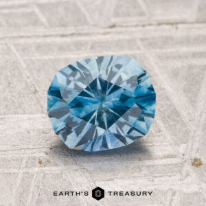 A faceted 1.58-carat heated Montana sapphire rests on a textured gray surface; the "EARTH'S TREASURY" logo and text appear at the bottom.
