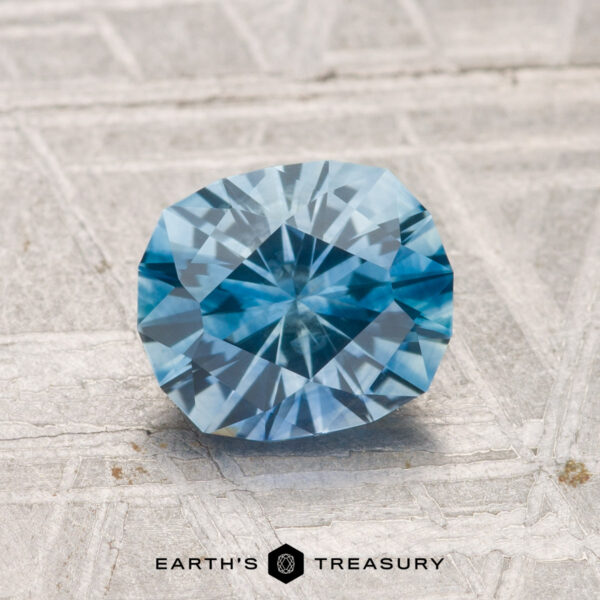 1.58-Carat Montana Sapphire (Heated) A faceted 1.58-carat heated Montana sapphire rests on a textured gray surface; the "EARTH'S TREASURY" logo and text appear at the bottom.