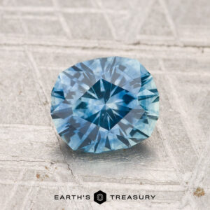 A faceted, cushion-cut 1.48-carat Montana sapphire (heated) in blue, displayed on a textured light gray surface with the "Earth's Treasury" logo at the bottom.