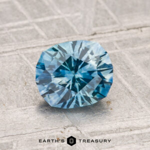 A 1.44-carat heated Montana sapphire with a blue, round geometric cut is displayed on a textured gray surface, with the "Earth's Treasury" logo and text at the bottom.
