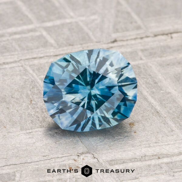 1.44-Carat Montana Sapphire (Heated) A 1.44-carat heated Montana sapphire with a blue, round geometric cut is displayed on a textured gray surface, with the "Earth's Treasury" logo and text at the bottom.