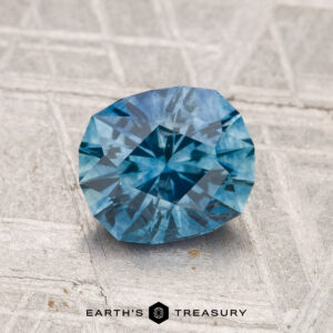 A 1.42-carat heated Montana sapphire is displayed on a textured gray surface with "Earth's Treasury" at the bottom.