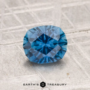 A 1.34-carat heated Montana sapphire, blue and faceted, rests on a textured light gray surface with "Earth's Treasury" displayed at the bottom.