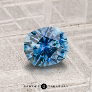 A 1.33-carat heated Montana sapphire with a geometric cut rests on a textured gray surface. The "Earth's Treasury" logo appears at the bottom of the image.